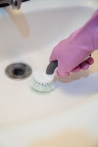 Person pouring baking soda and vinegar into a kitchen sink drain with a kettle of hot water nearby.