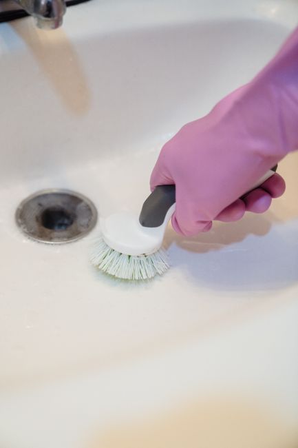 Person pouring baking soda and vinegar into a kitchen sink drain with a kettle of hot water nearby.
