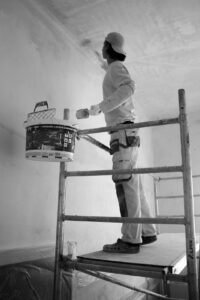 A person in work clothes carefully painting a residential room wall with a roller and tray, with painter’s tape on the trim.