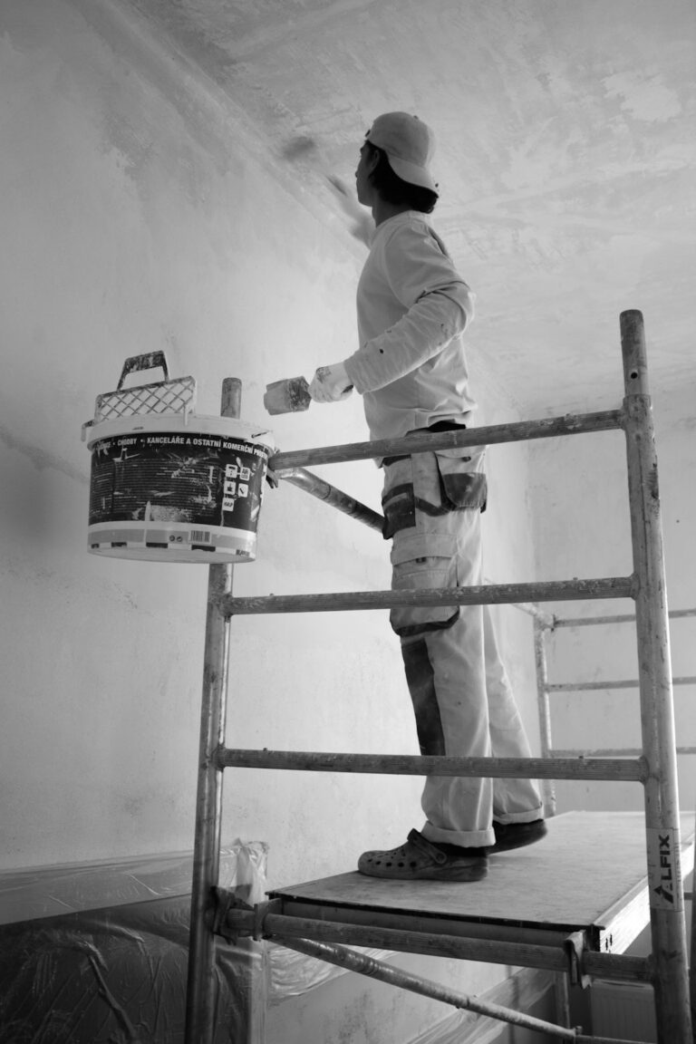 A person in work clothes carefully painting a residential room wall with a roller and tray, with painter’s tape on the trim.